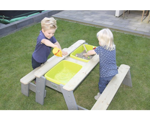 Zwei Kinder spielen an einem grauen Picknicktisch mit Sand und Wasser in grünen Behältern.