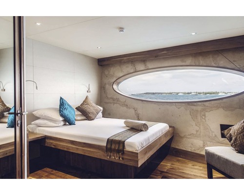 Helles Schlafzimmer mit Bett, Kissen und ovalem Fenster mit Meerblick