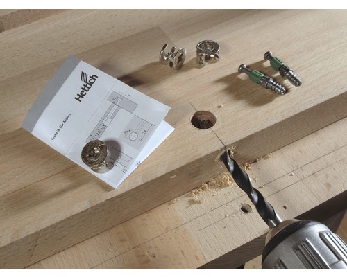 Bohrmaschine bohrt Loch in Holzbrett neben Möbelverbindern und Hettich Logo