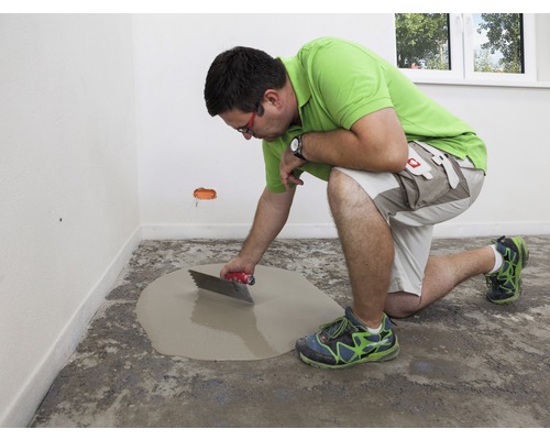 Handwerker, der Bodenausgleichsmasse mit einer Glättkelle in einem Raum verteilt.
