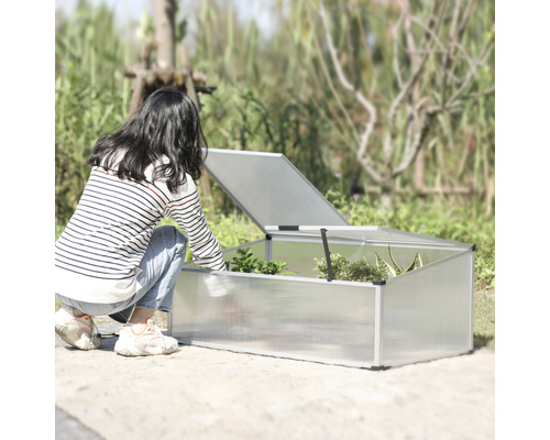 Frau beugt sich zu einem Frühbeetkasten mit geöffnetem Deckel im Garten.