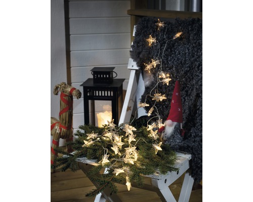 LED Lichterkette Konstsmide Acryl Sterne 3,9 m 40 LEDs Lichtfarbe warmweiß batteriebetrieben inkl. Timer Dekorative Szene mit Sternenlichterkette, Laterne und Zwerg auf einem Hocker.