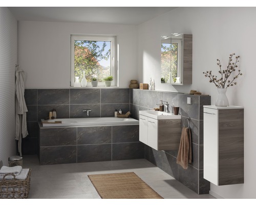 Badezimmer mit Badewanne, Waschtisch, Spiegelschrank und Hochschrank in hellem und dunklem Holzdekor sowie grauen Fliesen.