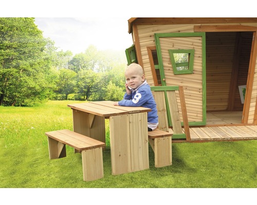 Kind sitzt am Holztisch mit Bank vor einem Holzspielhaus im Garten