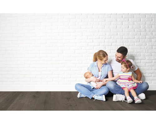 Familie sitzt auf einem dunklen Holzboden vor einer hellen Backsteinwand.