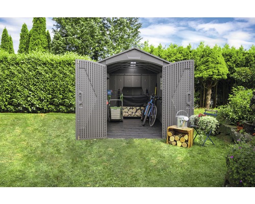 Garten mit offenem Geräteschuppen, in dem Gartengeräte, ein Fahrrad und Holz gelagert werden.