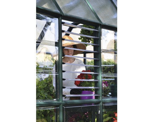 Eine Frau mit Hut gärtnert in einem Gewächshaus mit Lamellenfenstern und Blumen.