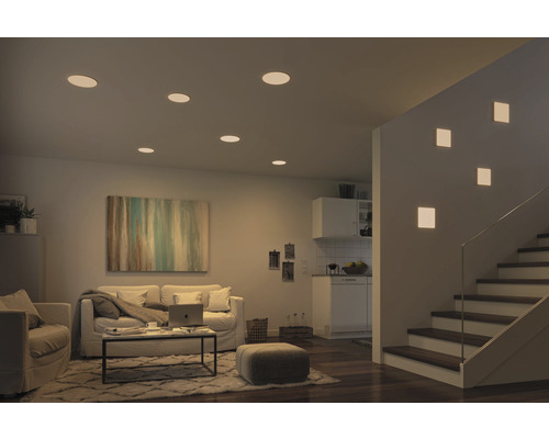 Wohnzimmer mit Deckeneinbaustrahlern und Wandleuchten neben einer Treppe.