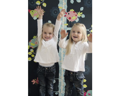 Zwei Mädchen stehen vor einer Kindermesslatte mit Vogelmotiven an einer dunklen Wand mit Blumenmuster.