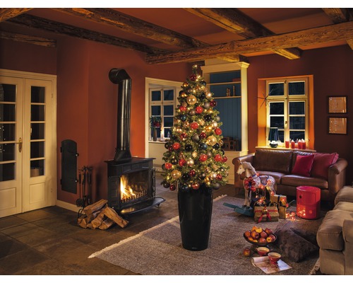 Weihnachtliche Wohnrauminszenierung mit Kamin, Couch und geschmücktem Weihnachtsbaum im Pflanzgefäß.