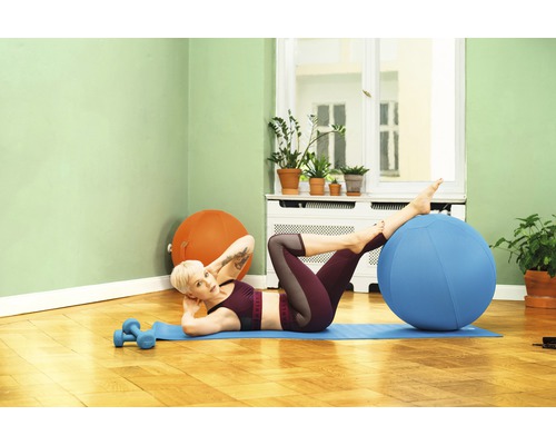 Frau macht Übungen mit einem Gymnastikball auf einer Matte im Wohnzimmer.