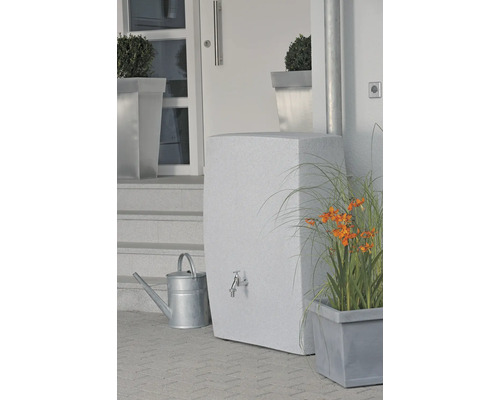Wassertank mit Wasserhahn, Gießkanne und Bepflanzung vor einem Haus