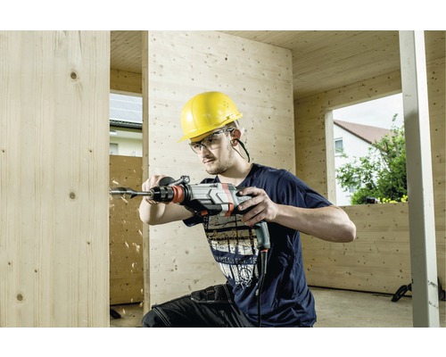 Ein Mann bohrt mit einer Bohrmaschine ein Loch in ein Holzbrett. Er trägt einen Schutzhelm und eine Schutzbrille.