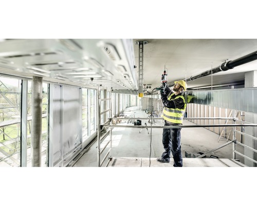 Ein Bauarbeiter auf einem Gerüst arbeitet mit einem Bohrhammer an einer Deckeninstallation.