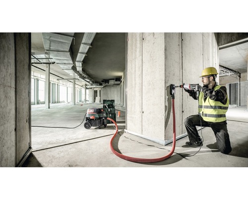 Ein Bauarbeiter bohrt mit einem Bohrhammer ein Loch in eine Betonwand, Staubabsaugung ist angeschlossen.