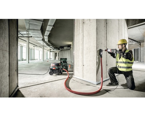 Ein Bauarbeiter bohrt mit einer Bohrmaschine ein Loch in eine Betonwand. Ein Industriestaubsauger und ein Absaugadapter sind ebenfalls vorhanden.