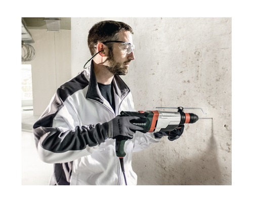 Ein Mann bohrt mit einem Metabo Bohrhammer ein Loch in eine Betonwand. Er trägt dabei eine Schutzbrille, Ohrenschützer und Handschuhe.