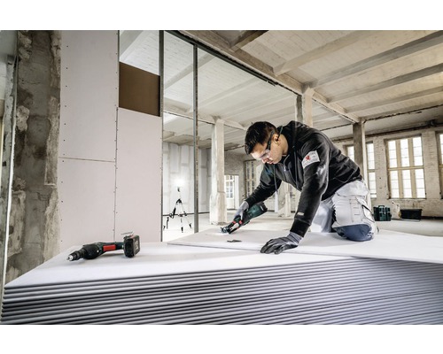 Ein Handwerker schneidet eine Gipskartonplatte mit einem Spezialwerkzeug in einem Rohbau, wobei andere Werkzeuge bereitliegen.