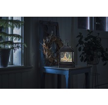 Dekorative Schneekugellaterne mit Winterdorf-Motiv auf einem Tisch, umgeben von Pflanzen und einem Kranz.