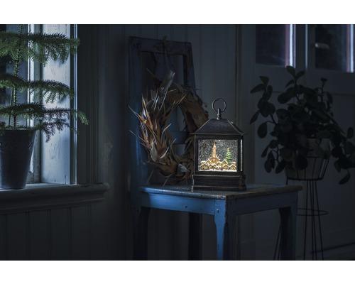 Dekorative Schneekugellaterne mit Winterdorf-Motiv auf einem Tisch, umgeben von Pflanzen und einem Kranz.