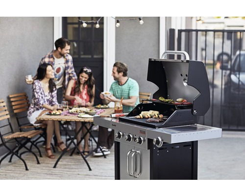 Personen grillen auf einer Terrasse mit einem Gasgrill und genießen eine Mahlzeit am Tisch.