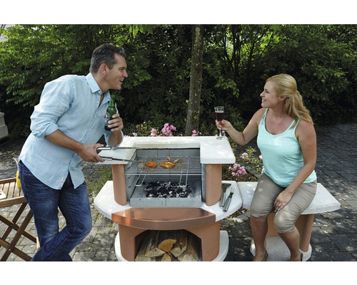 Buschbeck Grillkamin-Bar Bologna 65 x 110 x 93 cm Beton weiß Mann und Frau grillen im Garten mit einem gemauerten Grill.