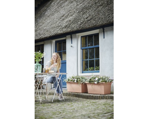 Eine Frau sitzt an einem Gartentisch vor einem Haus mit Blumenkästen und Reetdach.