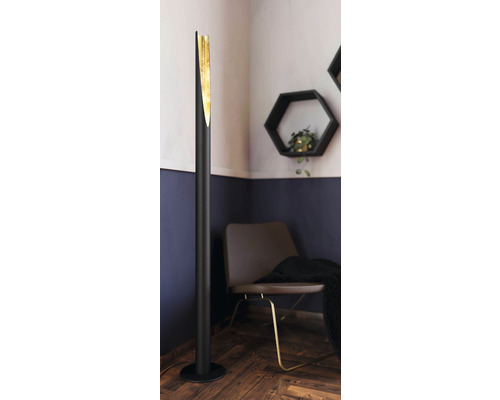 LED Stehlampe 340 lm 3000 K HxØ 137x6 cm Barbotto schwarz/gold mit Fußschalter Schwarze Stehlampe mit goldenem Detail neben einem Stuhl und einem sechseckigen Wandregal.