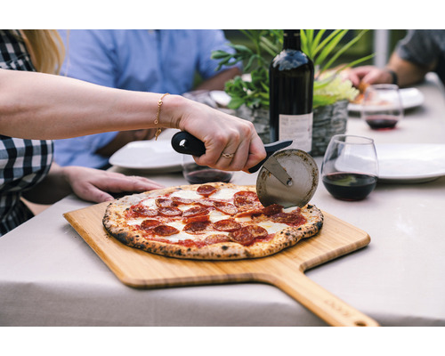 Szene einer Person, die eine Pizza mit einem Pizzaschneider auf einem Holzbrett schneidet, mit einer Weinflasche und Gläsern im Hintergrund, auf einem Tisch im Freien