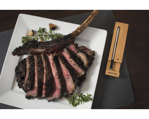 Tomahawk Steak auf einem Teller mit Kräutern und Knoblauch, zusammen mit einem Fleischthermometer