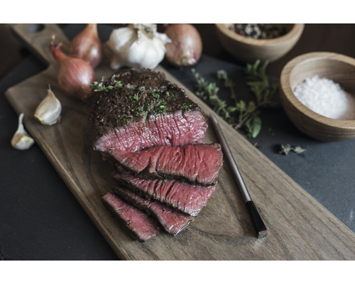 Auf einem Holzbrett liegt ein aufgeschnittenes Stück Fleisch, garniert mit Kräutern, zusammen mit Zwiebeln, Knoblauch, Pfeffer und Salz.