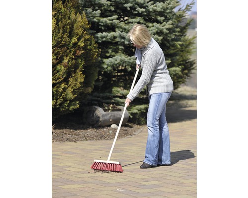 Frau kehrt mit einem Straßenbesen eine gepflasterte Fläche im Garten.