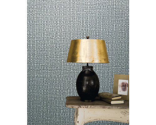 Nahaufnahme einer Wand mit Textiltapete neben einem Tisch mit Lampe, Büchern und Fotorahmen.