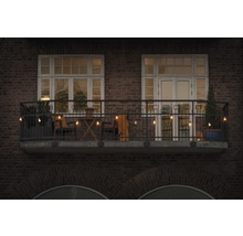 Dekorierter Balkon mit Lichterkette, Stühlen, Tisch und Pflanzen vor einem Backsteingebäude.