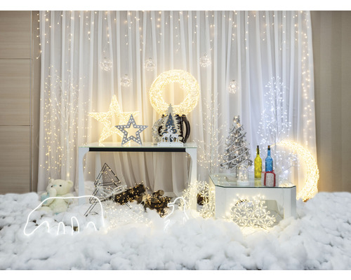 Weihnachtliche Dekoration mit Lichtern, Sternen und Ornamenten auf Tischen vor einem weißen Vorhang.