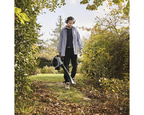 Ein Mann mit Gehörschutz und Handschuhen benutzt einen Laubsauger im Garten.