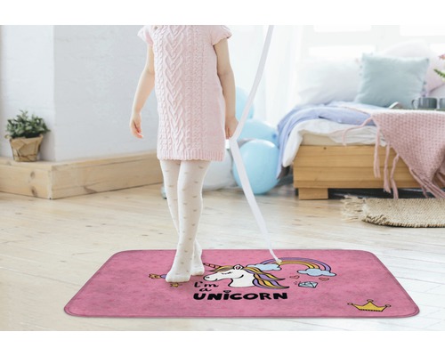 Mädchen steht auf einem pinkfarbenen Teppich mit Einhornmotiv in einem hellen Raum.
