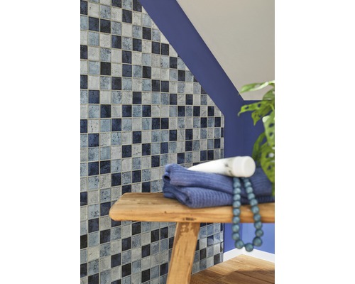 Badezimmerwand mit Mosaikfliesen in verschiedenen Blautönen, Handtuch und Dekoration auf einem Holzhocker.