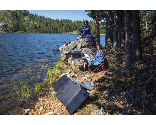 Szene am See mit tragbarem Solarmodul, Powerstation, Tisch und Campingstühlen für den Outdoor-Einsatz.