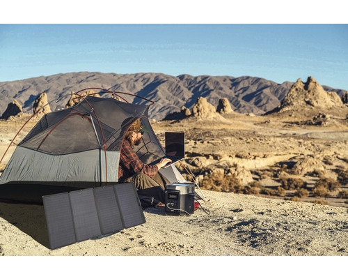 Mann arbeitet im Zelt mit Laptop, Stromgenerator und Solarmodul in Berglandschaft.