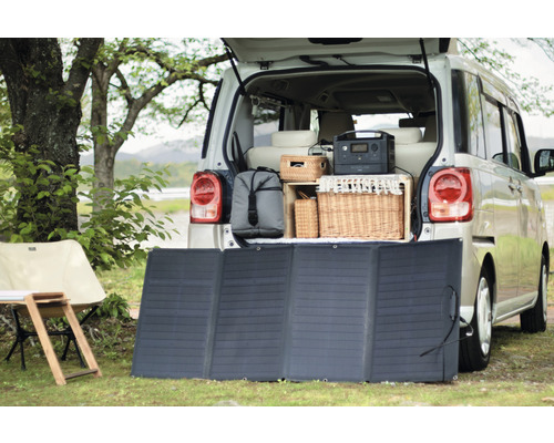 Szene mit einem Solarmodul vor einem geöffneten Kofferraum eines Autos mit Campingausrüstung.