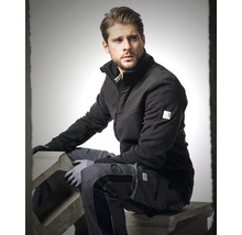 Mann in Arbeitskleidung mit Fleecejacke und Arbeitshose sitzt auf einem Betonblock