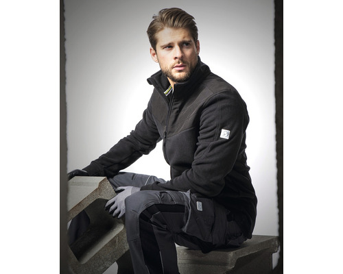 Mann in Arbeitskleidung mit Fleecejacke und Arbeitshose sitzt auf einem Betonblock