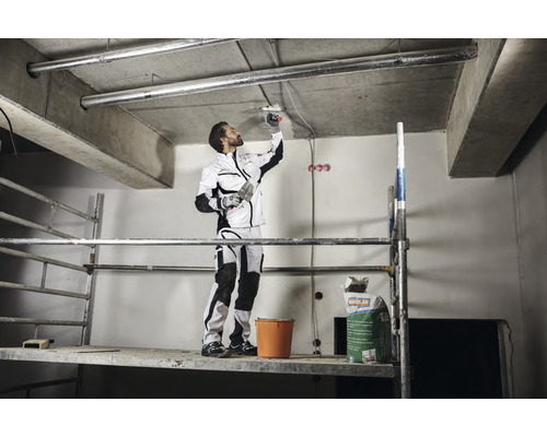 Handwerker auf einem Gerüst, der mit einer Spachtel Deckenputz aufträgt. Im Hintergrund befindet sich ein Sack Putz.