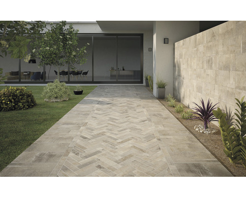 Außenbereich mit Fliesenweg im Fischgrätmuster, Rasenfläche und bepflanzten Beeten.