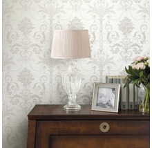Dekoratives Interieur mit gemusterter Tapete, Lampe, Bilderrahmen, Büchern und Blumen auf einem Schrank.