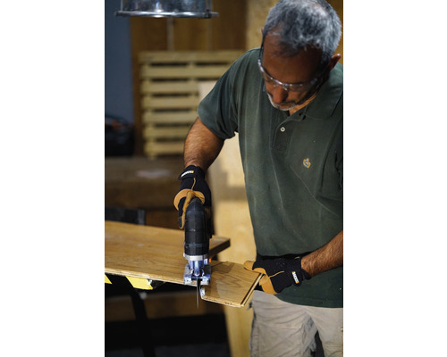 Ein Mann benutzt eine Stichsäge, um ein Stück Holz zu schneiden. Er trägt dabei Schutzbrille und Handschuhe.