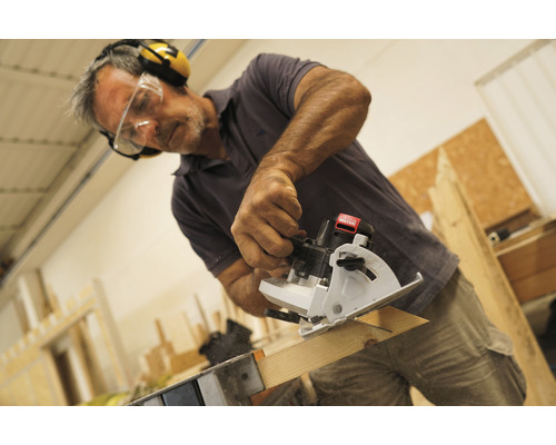 Mann sägt Holzbrett mit einer Mini-Handkreissäge, während er Schutzbrille und Gehörschutz trägt.