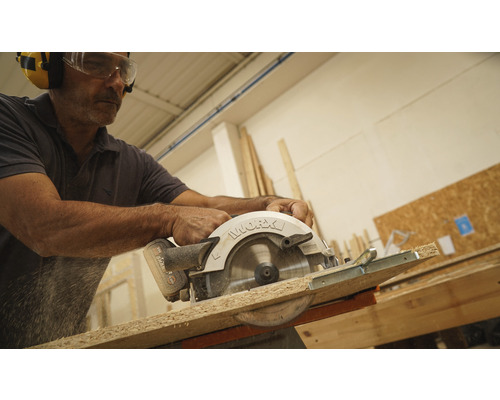 Mann sägt mit einer Handkreissäge ein Holzbrett, wobei er eine Schutzbrille und Ohrenschützer trägt.