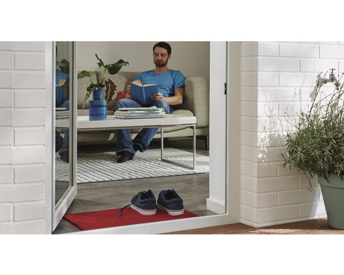 Ein Mann liest ein Buch in einem Wohnzimmer, das durch eine offene Tür mit einer Fußmatte und Schuhen zu sehen ist.